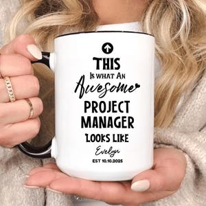 Può includere: Tazza in ceramica bianca con manico e bordo neri. La tazza presenta la scritta "THIS Is WHAT AN Awesome PROJECT MANAGER LOOKS LIKE Evelyn EST 10.10.2025" in nero. La tazza è tenuta da una persona.