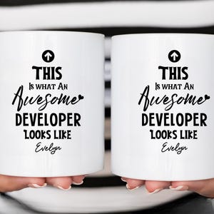 Op de afbeelding: Twee witte keramische mokken met zwarte tekst met de tekst "THIS IS WHAT AN Awesome DEVELOPER LOOKS LIKE" met de naam "Evelyn" eronder. Elke mok heeft een zwarte pijl omhoog.