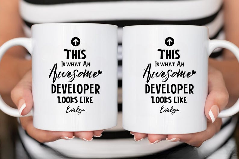 Op de afbeelding: Twee witte keramische mokken met zwarte tekst met de tekst "THIS IS WHAT AN Awesome DEVELOPER LOOKS LIKE" met de naam "Evelyn" eronder. Elke mok heeft een zwarte pijl omhoog.