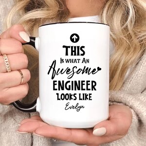 May include: White ceramic mug with a black handle and rim. The mug features the text "THIS Is WHAT An Awesome ENGINEER LOOKS LIKE Evelyn" in black script and a black arrow. The mug is held by a person.