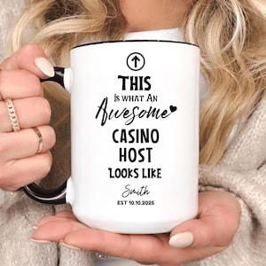 Pode incluir: Caneca de cerâmica branca com alça e borda pretas. A caneca apresenta o texto "THIS Is WHAT AN Awesome CASINO HOST LOOKS LIKE" com um nome e uma data abaixo. A caneca é segurada por uma pessoa.