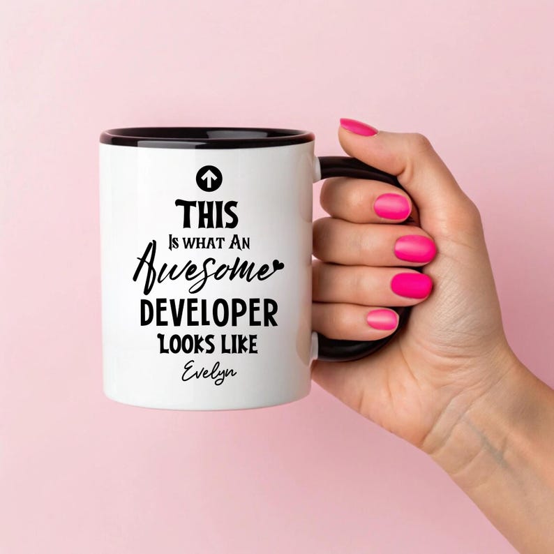 Op de afbeelding: Witte keramische mok met een zwarte binnenkant en handvat. De mok heeft de tekst "THIS IS WHAT AN Awesome DEVELOPER LOOKS LIKE Evelyn" in zwarte letters. De mok wordt vastgehouden door een hand met roze nagellak tegen een roze achtergrond.