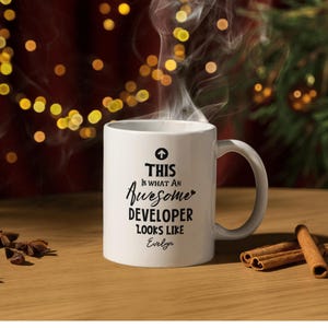 Op de afbeelding: Een witte keramische mok met de tekst "THIS IS WHAT AN AWESOME DEVELOPER LOOKS LIKE" in het zwart. De mok heeft een oor en is gevuld met een warme drank, met stoom die opstijgt. De naam "Evelyn" staat eronder.