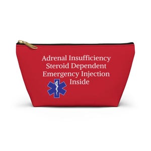 Pode incluir: Uma bolsa vermelha com zíper dourado e acabamento preto. A bolsa apresenta texto branco que diz "Adrenal Insufficiency Steroid Dependent Emergency Injection Inside" com um símbolo médico azul.