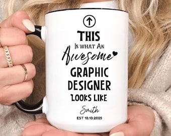 Op de afbeelding: Witte keramische mok met een zwart handvat en rand. De mok heeft de tekst "THIS IS WHAT AN Awesome GRAPHIC DESIGNER LOOKS LIKE Smith EST 10.10.2025" in zwart schrift en een zwart pijlsymbool.