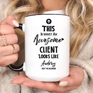 Puede incluir: Taza de cerámica blanca con asa y borde negros. La taza presenta el texto "THIS IS WHAT AN Awesome CLIENT LOOKS LIKE Audrey EST 10.10.2025" en escritura negra y una flecha negra. La taza es sostenida por una persona.