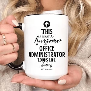 Puede incluir: Taza de cerámica blanca con asa y borde negros. La taza presenta el texto "THIS IS WHAT AN Awesome OFFICE ADMINISTRATOR LOOKS LIKE Audrey EST 10.10.2025" en letras negras con un gráfico de flecha.