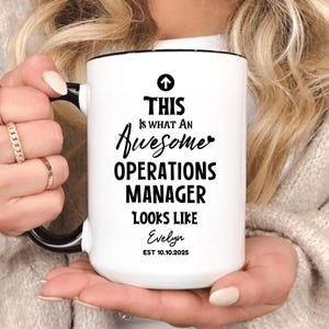Puede incluir: Taza de cerámica blanca con asa y borde negros. La taza presenta el texto "THIS Is WHAT An Awesome OPERATIONS MANAGER LOOKS LIKE Evelyn EST 10.10.2025" en letras negras.