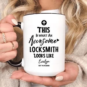 Puede incluir: Taza de cerámica blanca con asa y borde negros. La taza presenta el texto "THIS Is WHAT An Awesome LOCKSMITH LOOKS LIKE" en negro, con el nombre "Evelyn" y la fecha "EST 10.10.2025" debajo. La taza es sostenida por una persona.