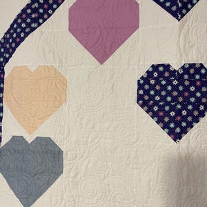 May include: A white quilt with a patchwork design featuring hearts in various colors and patterns. The hearts are made of different fabrics, including a pink heart with a floral pattern, a light blue heart with a textured pattern, and a peach heart with a solid color.
