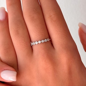 Puede incluir: Un delicado anillo de plata con una fila de pequeñas piedras redondas y transparentes. El anillo está en el dedo de una persona.