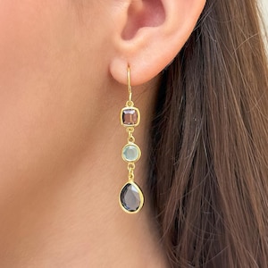 May include: Gold drop earrings featuring three gemstones: a square, a round, and a teardrop. The gemstones are reddish-brown, light blue-green, and dark grey. The earrings are shown on a person's ear.