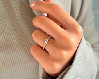 May include: A gold ring with a single, clear, rectangular-shaped stone set in the center. The ring is surrounded by smaller, clear stones.