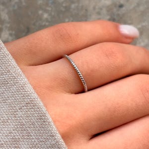 May include: A thin silver ring with a braided design. The ring is worn on a person's finger.