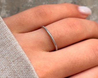 May include: A thin silver ring with a braided design. The ring is worn on a person's finger.