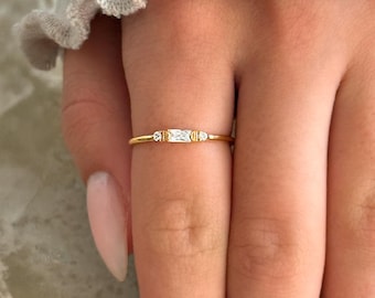 May include: A delicate gold ring with a rectangular clear stone and two smaller clear stones on either side.