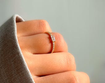 May include: A gold ring with a single, clear, rectangular-shaped stone set in the center. The band is adorned with small, sparkling stones.