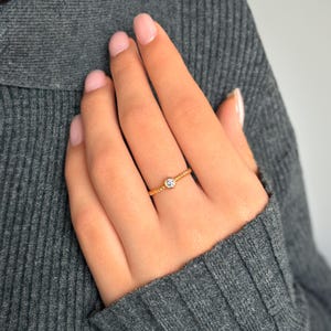 Könnte beinhalten: Ein zarter Goldring mit einem kleinen, runden, klaren Edelstein, der in der Mitte sitzt. Die Band ist verdreht und hat einen dezenten Glanz.