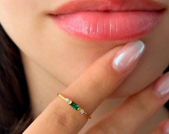 May include: A delicate gold ring featuring a rectangular emerald-cut green gemstone, flanked by two small, round, clear gemstones. The ring is displayed on a finger, with a soft pink lip and skin in the background.