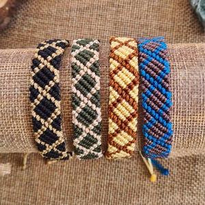 Può includere: Quattro braccialetti macramè fatti a mano in nero e oro, verde e beige, marrone e giallo, e blu e marrone. I braccialetti sono esposti su una superficie di juta, con fette di legno e cristalli sullo sfondo.