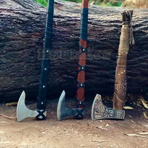 May include: Three axes standing upright. The first has a black handle with leather wrapping. The second has a dark wood handle with black leather lacing. The third has a light wood handle with twine wrapping and an etched metal blade.