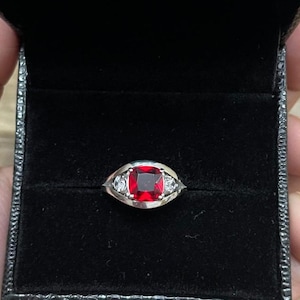 May include: A silver ring with a large, square, red gemstone at the center, flanked by two smaller, clear stones. The ring is displayed in a black jewelry box with a textured exterior.
