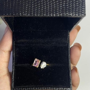 Op de afbeelding: Een gouden ring met een roze rechthoekige edelsteen en een witte peervormige edelsteen, gepresenteerd in een open zwarte sieradendoos met textuur. De binnenkant van de doos is zwart fluweel. De doos heeft een gouden logo.