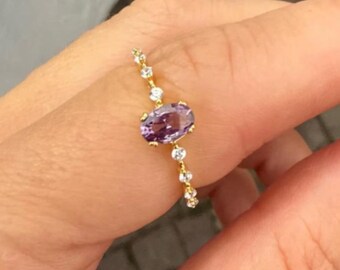 Puede incluir: Un anillo de oro con una piedra preciosa ovalada púrpura en el centro, flanqueada por una línea de piedras preciosas más pequeñas y claras. El anillo se muestra en un dedo, destacando su diseño delicado y elegante.