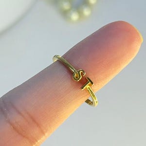Puede incluir: Un anillo dorado con las letras "S" y "T". El anillo es abierto y ajustable, con las letras formando el elemento principal del diseño. El anillo está en un dedo, con un fondo borroso.