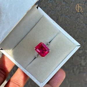 Pode incluir: Um anel com uma grande gema vermelha quadrada brilhante engastada em uma banda prateada. O anel está em uma caixa de anel branca.
