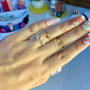 Puede incluir: Un anillo de oro con una piedra preciosa cuadrada transparente y piedras preciosas transparentes más pequeñas en la banda. El anillo se lleva en un dedo con esmalte de uñas rosa y blanco adornado con diamantes de imitación.