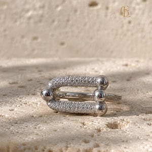 May include: A silver-toned ring featuring a U-shaped centerpiece adorned with small, sparkling clear stones. The design includes three smooth, polished spherical accents at the ends of the U-shape, creating a distinct, modern, and geometric aesthetic.