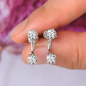 May include: A pair of silver earrings featuring two clear, round gemstones. The earrings have a post and a dangling design, with a gemstone at the top and bottom. The gemstones sparkle, reflecting light. The earrings are held by a hand.