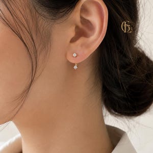 May include: A gold earring featuring two small, clear, round-cut stones in a prong setting. One stone sits directly on the earlobe, while the second stone hangs slightly below it, connected by a thin gold bar. A small logo is visible in the corner.