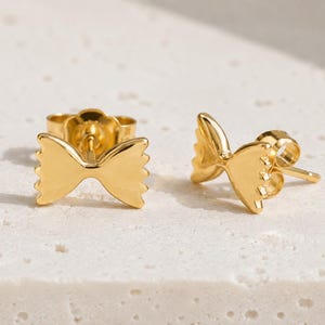 Pode incluir: Um par de brincos de tarracha dourados em formato de massa farfalle. Cada brinco apresenta um franzido central com bordas serrilhadas em ambos os lados, imitando a aparência de uma gravata borboleta. Incluem fechos de pino padrão.