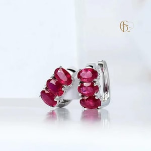 Könnte beinhalten: Ein Paar silberne Creolen, jeweils besetzt mit drei oval geschliffenen, leuchtend roten Edelsteinen. Die Ohrringe sind vor einem hellweißen Hintergrund platziert, was das elegante Design und die Farbe des Schmucks hervorhebt.