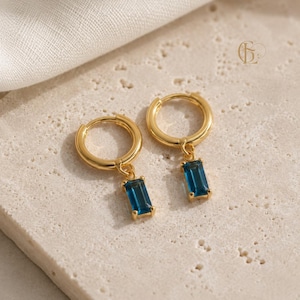 Può includere: Un paio di orecchini a cerchio dorati, ciascuno con una pietra preziosa blu rettangolare a taglio baguette pendente. Gli orecchini hanno una finitura metallica lucida e una chiusura a cerniera sicura. Un piccolo logo è visibile in alto a destra.