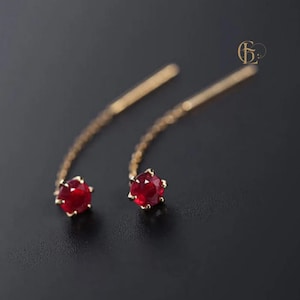 Könnte beinhalten: Ein Paar goldene Ohrringe mit einem hängenden Design. Jeder Ohrring hat einen leuchtend roten Edelstein am unteren Ende, der in einer goldenen Lünette gefasst ist. Die Ohrringe haben eine gebogene Form mit einem kettenartigen Detail, das zum Edelstein führt.