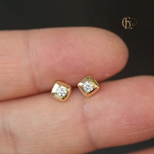Könnte beinhalten: Ein Paar goldfarbene quadratische Ohrstecker, jeweils mit einem kleinen, funkelnden Diamanten in der Mitte. Die Ohrringe sind vor einem unscharfen Hintergrund platziert.