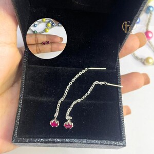 May include: Silver threader earrings with a small, round, red gemstone at the end of each chain. The earrings are presented in a black jewellery box. A close-up shows a pair of small, red gemstone earrings.
