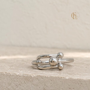 Puede incluir: Un anillo de tono plateado pulido con un diseño en forma de U y tres acentos esféricos. El centro de la forma en U sostiene una esfera más grande, mientras que dos esferas más pequeñas se sitúan en los extremos abiertos de la banda metálica.