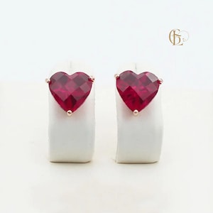 Könnte beinhalten: Ein Paar herzförmige, rubinrote Ohrstecker. Die Ohrringe sind in Roségold gefasst und werden auf weißen Ständern vor weißem Hintergrund präsentiert. Die Ohrringe sind ein romantisches Schmuckstück.