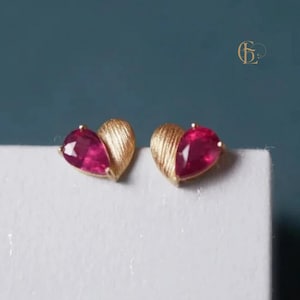 Puede incluir: Un par de pendientes de botón en forma de corazón. Cada pendiente presenta una media forma de corazón dorada texturizada y una piedra preciosa roja oscura en forma de lágrima. Los pendientes se muestran sobre una superficie blanca sobre un fondo verde azulado.