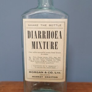 Peut inclure: Une bouteille de m&eacute;dicament en verre bleu clair transparent avec un corps rectangulaire et un col court. Une &eacute;tiquette en papier blanc est fix&eacute;e sur le devant, avec un texte noir indiquant SHAKE THE BOTTLE, DIARRHOEA MIXTURE, des instructions de dosage, des ingr&eacute;dients et MORGAN & CO. LTD. CHEMISTS MARKET DRAYTON.