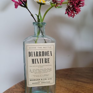 Peut inclure: Une bouteille en verre transparent au corps rectangulaire et au col &eacute;troit, contenant des fleurs roses et blanches. L'&eacute;tiquette indique SHAKE THE BOTTLE, DIARRHOEA MIXTURE, One table-spoonful every four hours in water, MORGAN & CO. LTD. CHEMISTS MARKET DRAYTON.