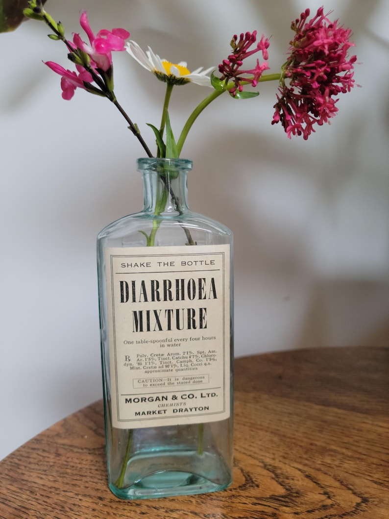 Peut inclure: Une bouteille en verre transparent au corps rectangulaire et au col &eacute;troit, contenant des fleurs roses et blanches. L'&eacute;tiquette indique SHAKE THE BOTTLE, DIARRHOEA MIXTURE, One table-spoonful every four hours in water, MORGAN & CO. LTD. CHEMISTS MARKET DRAYTON.