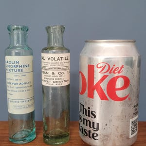 Peut inclure: Deux flacons de m&eacute;dicaments en verre transparent vintage avec des &eacute;tiquettes en papier blanc et une canette de soda Diet Coke en aluminium argent&eacute;. Le flacon de gauche est &eacute;tiquet&eacute; Kaolin Morphine Mixture et celui du milieu Sal Volatile.