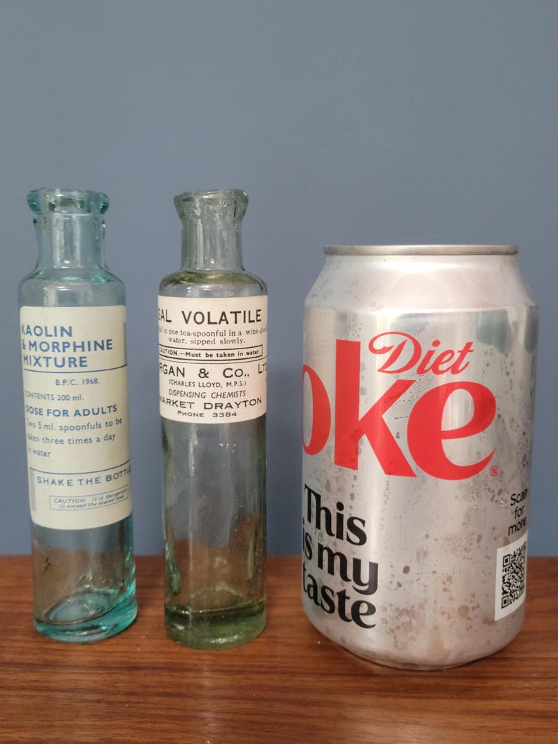 Peut inclure: Deux flacons de m&eacute;dicaments en verre transparent vintage avec des &eacute;tiquettes en papier blanc et une canette de soda Diet Coke en aluminium argent&eacute;. Le flacon de gauche est &eacute;tiquet&eacute; Kaolin Morphine Mixture et celui du milieu Sal Volatile.