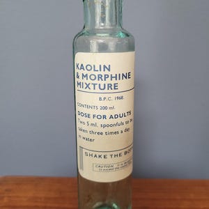 Peut inclure: Une bouteille de m&eacute;dicament en verre bleu clair transparent avec un corps cylindrique et un goulot &eacute;troit. L'&eacute;tiquette blanche porte le texte KAOLIN & MORPHINE MIXTURE, B.P.C. 1968, CONTENTS 200 ml, DOSE FOR ADULTS, SHAKE THE BOTTLE et CAUTION en bleu.