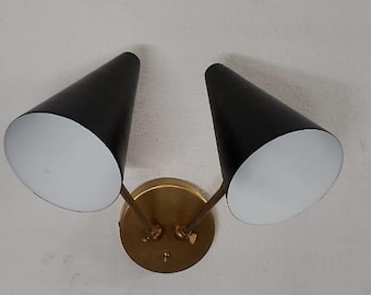 May include: A wall-mounted light fixture with two black cone-shaped shades. The interior of each shade is white. The fixture has a brass-colored base and arms. The light fixture is designed to be mounted on a wall.
